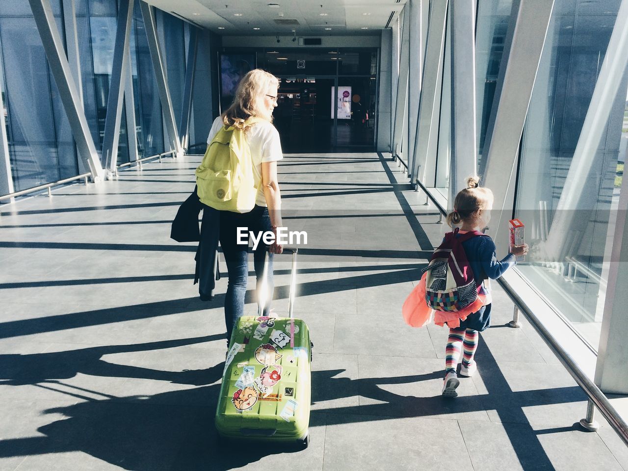 Rear view of mother pulling suitcase while walking with daughter at airport corridor