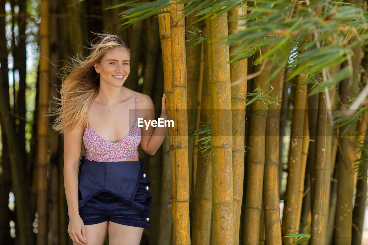 one person, smiling, women, adult, happiness, shorts, young adult, hairstyle, three quarter length, blond hair, leisure activity, front view, long hair, clothing, emotion, nature, standing, portrait, lifestyles, green, plant, fashion, tree, day, spring, trip, summer, vacation, cheerful, enjoyment, outdoors, holiday, yellow, land, casual clothing, person, carefree, looking at camera, travel, teeth, forest, smile, looking, dress, photo shoot, jungle, relaxation, rural scene, female