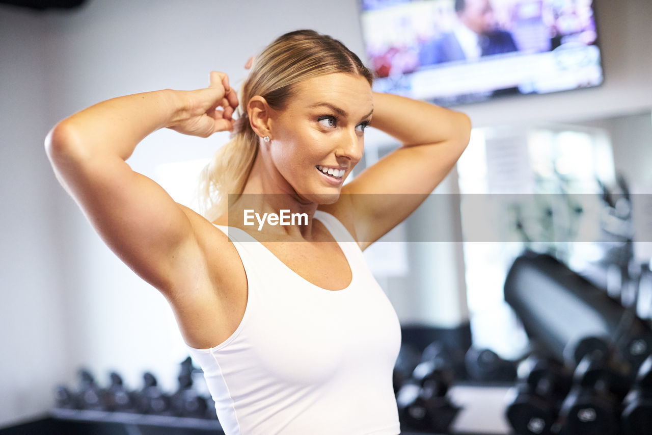 An indoor portrait of a blonde woman tying her hair back in the gym.