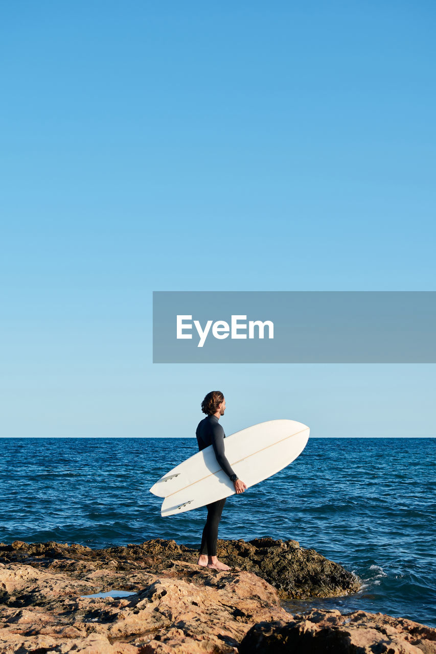 Side view of male surfer in wetsuit standing on rock near sea with surfboard and getting ready for surfing