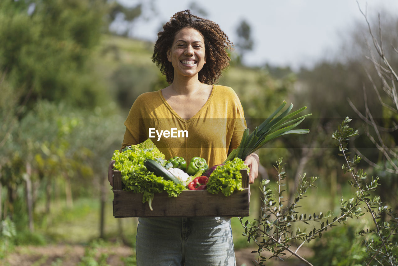 Smiling woman holding crate of fresh vegetables while standing in garden
