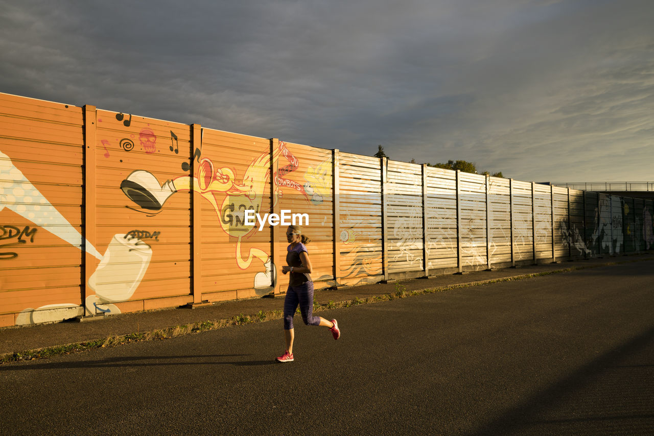 REAR VIEW OF WOMAN RUNNING ON FENCE AGAINST SKY