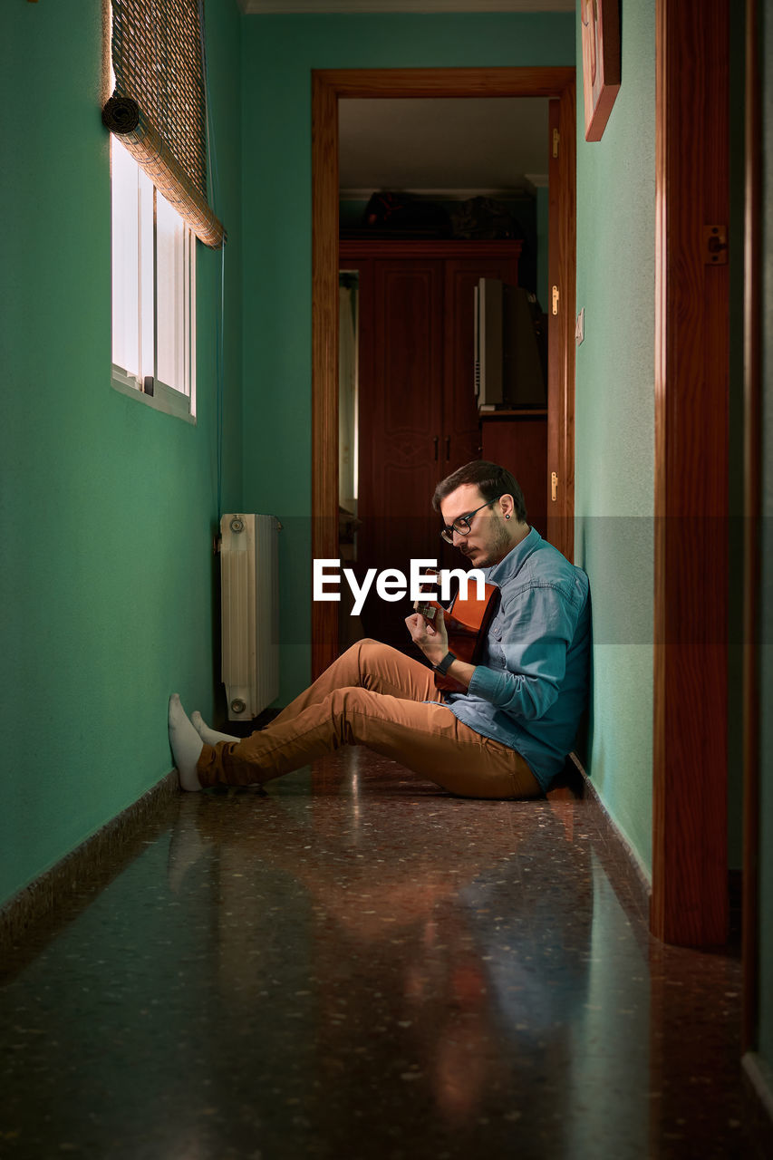 Young man plays guitar sitting in the hallway of his house