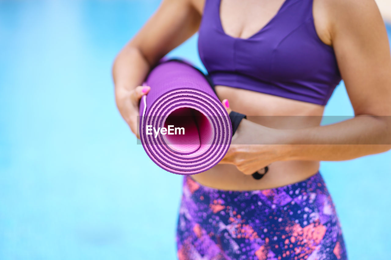 Horizontal view of unrecognizable woman holding a pink yoga mat with swimming pool background. 