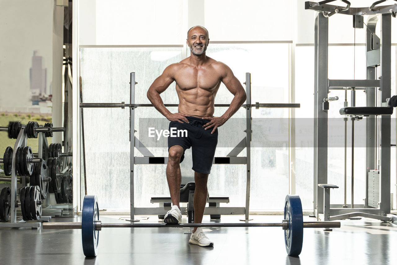 Full length of shirtless man with barbell standing in gym