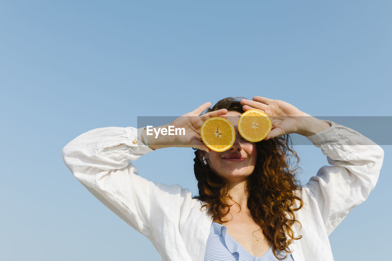 Smiling woman covering eyes with oranges on sunny day