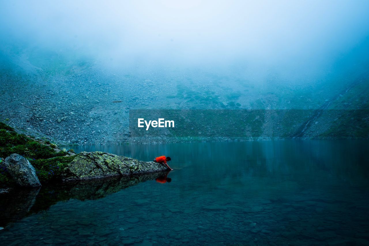 Side view of man on lakeshore against mountain during foggy weather