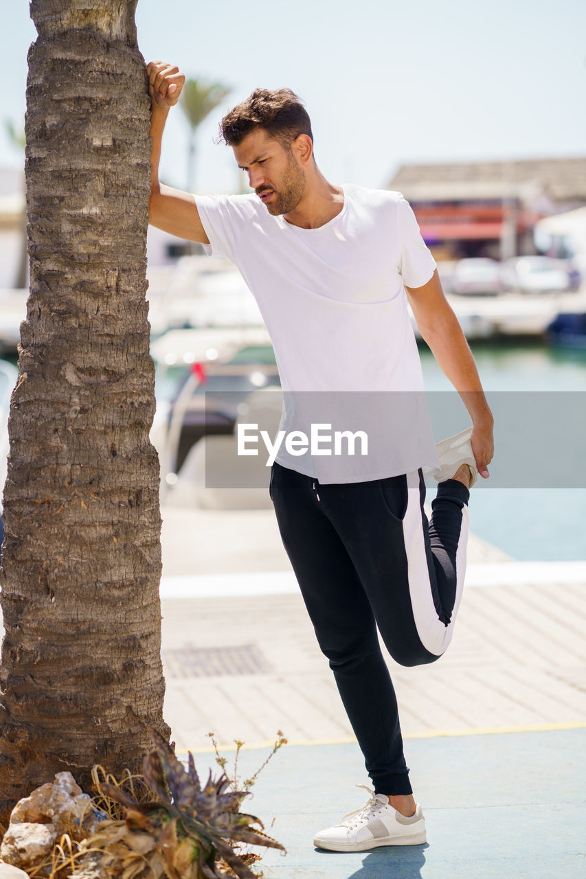 Full length of young man stretching by tree trunk at harbor