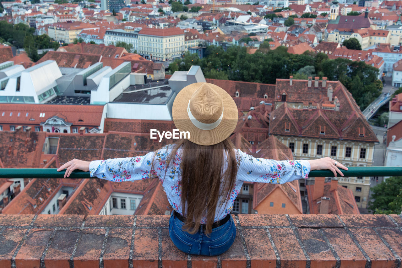 A girl with long hair in a hat sits on the edge of a tall building and looks at the city of graz. 