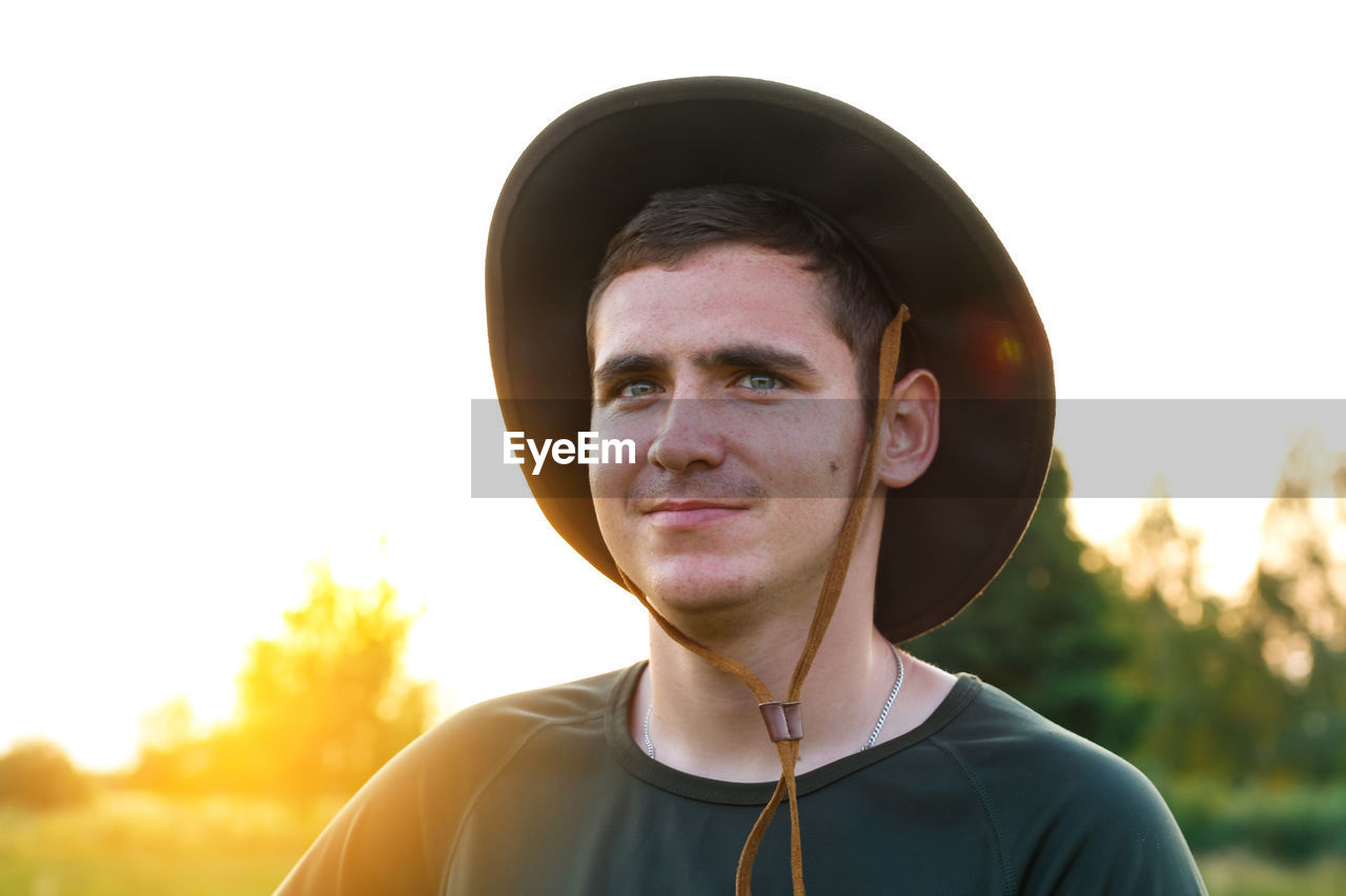 Young man smiling farmer in cowboy hat at agricultural field on sunset with sun flare. closeup 