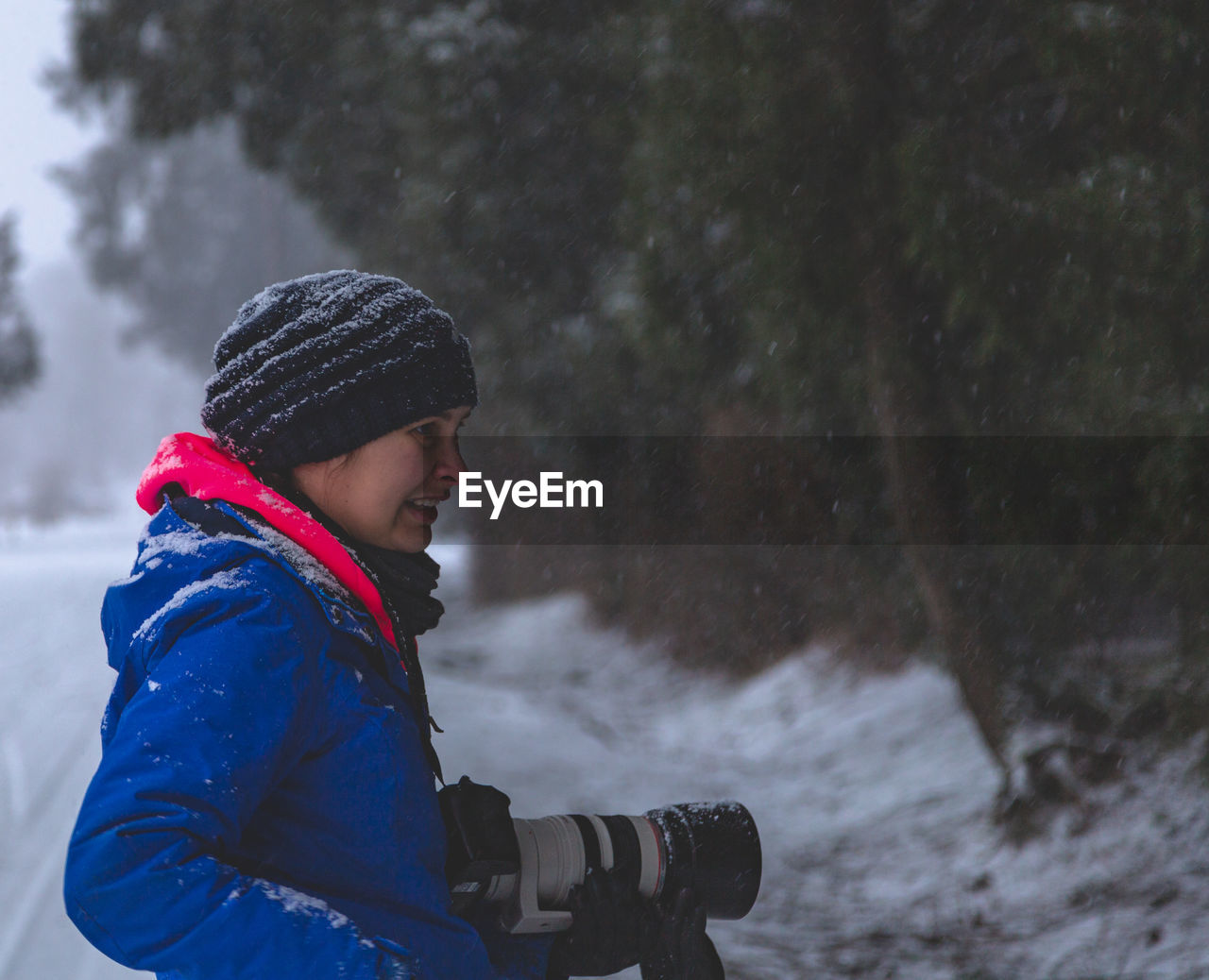 Side view of woman wearing warm clothing while holding digital camera in forest during snowfall