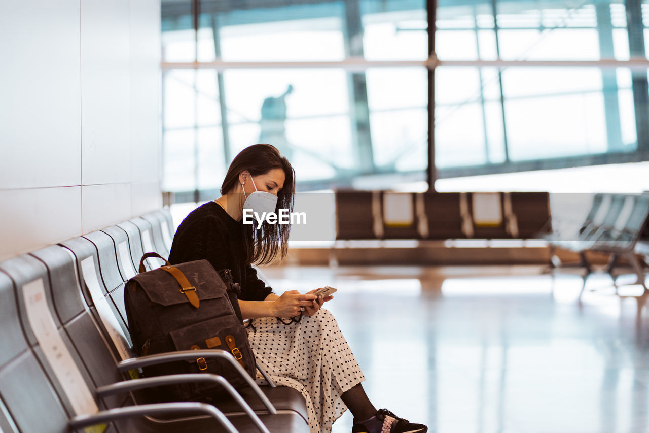 Woman wearing mask using mobile phone while sitting at airport