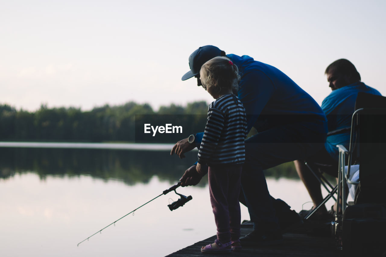 Rear view of daughter and father fishing in lake against sky