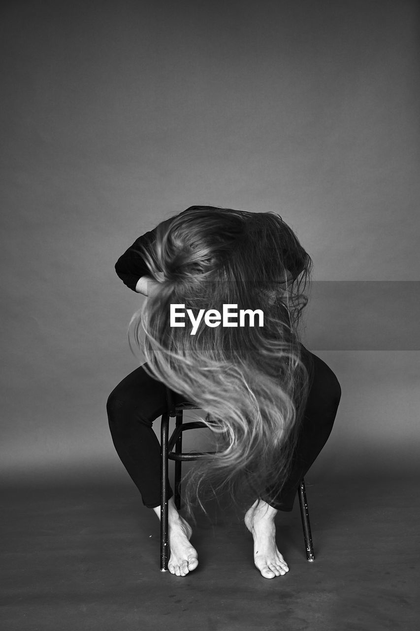 Woman covering her face by hair while sitting on chair against gray background