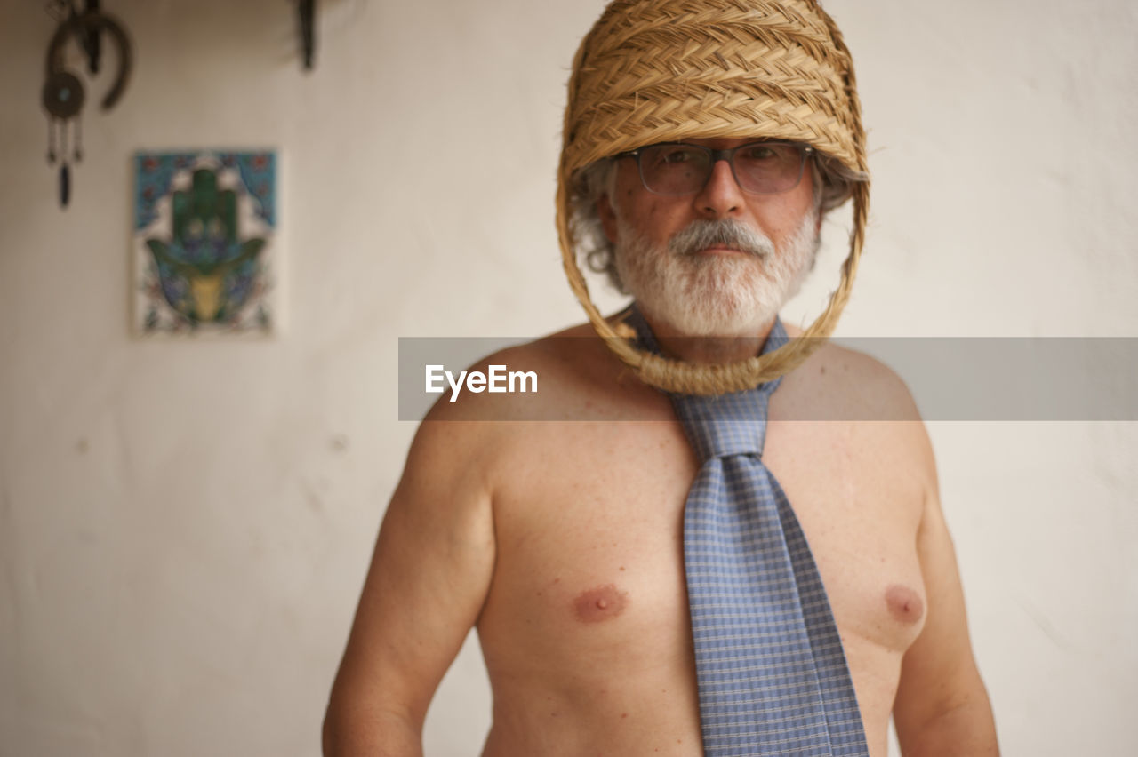 Portrait of shirtless man wearing basket while standing against wall