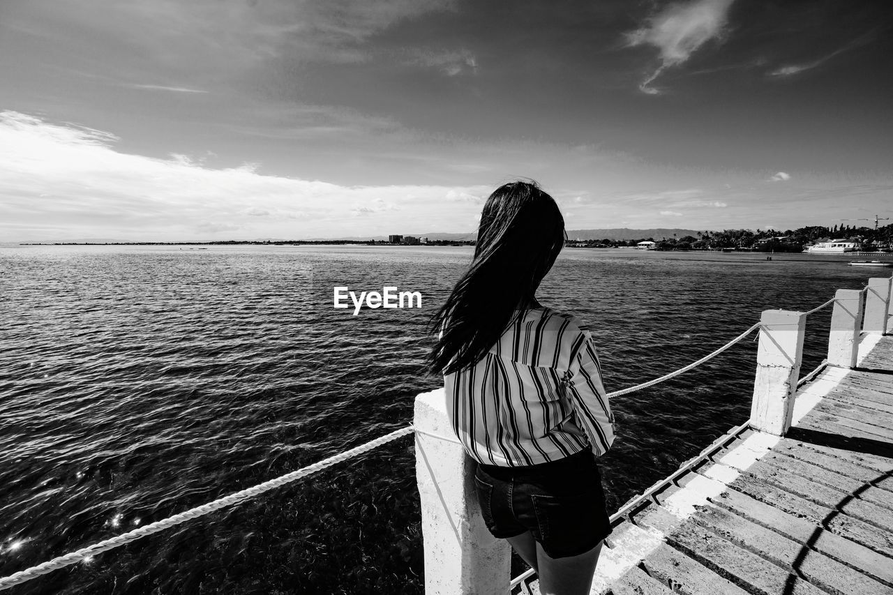 REAR VIEW OF WOMAN STANDING ON RAILING AGAINST SEA