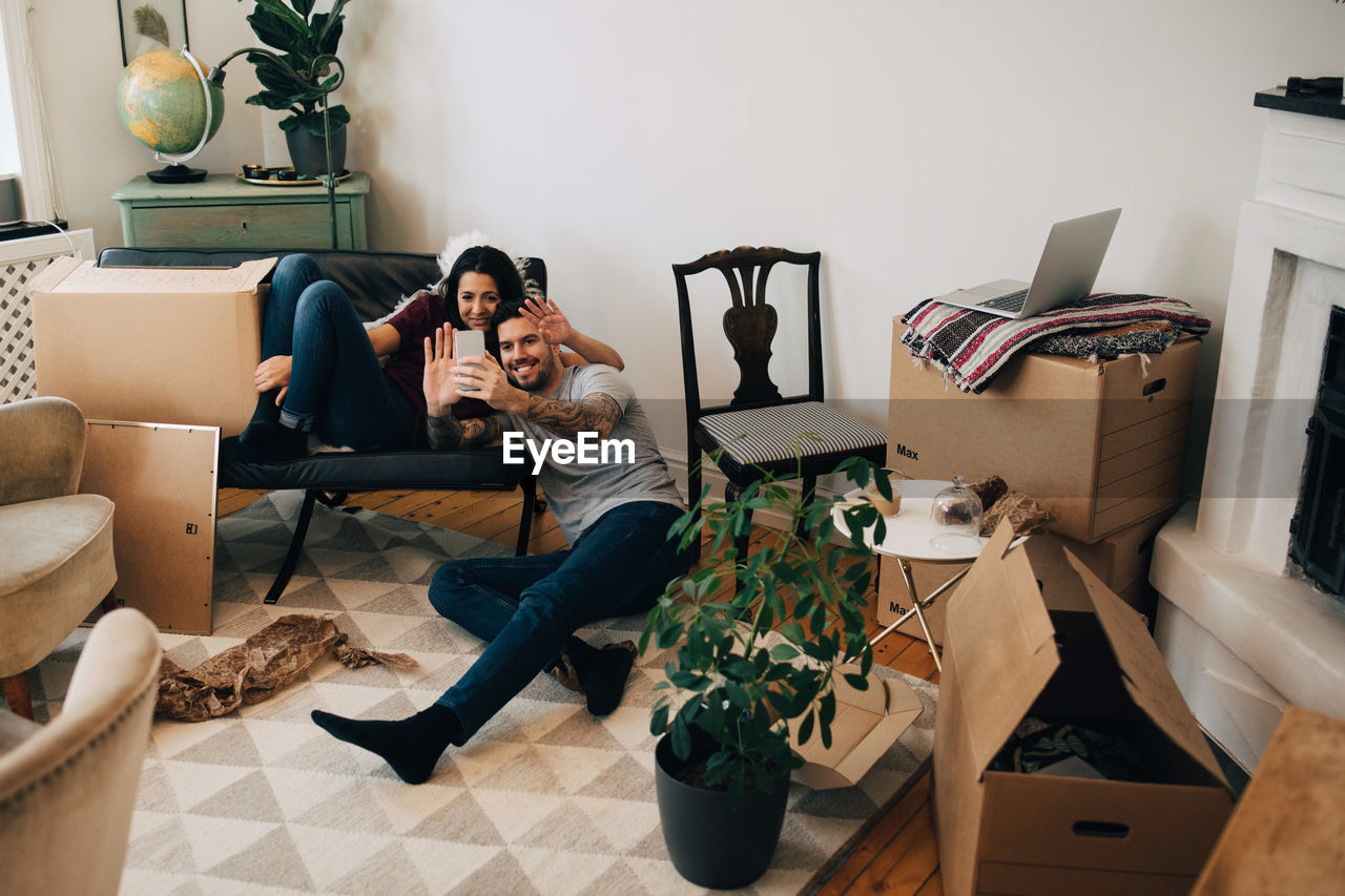 Smiling couple video calling through mobile phone while sitting in living room during relocation