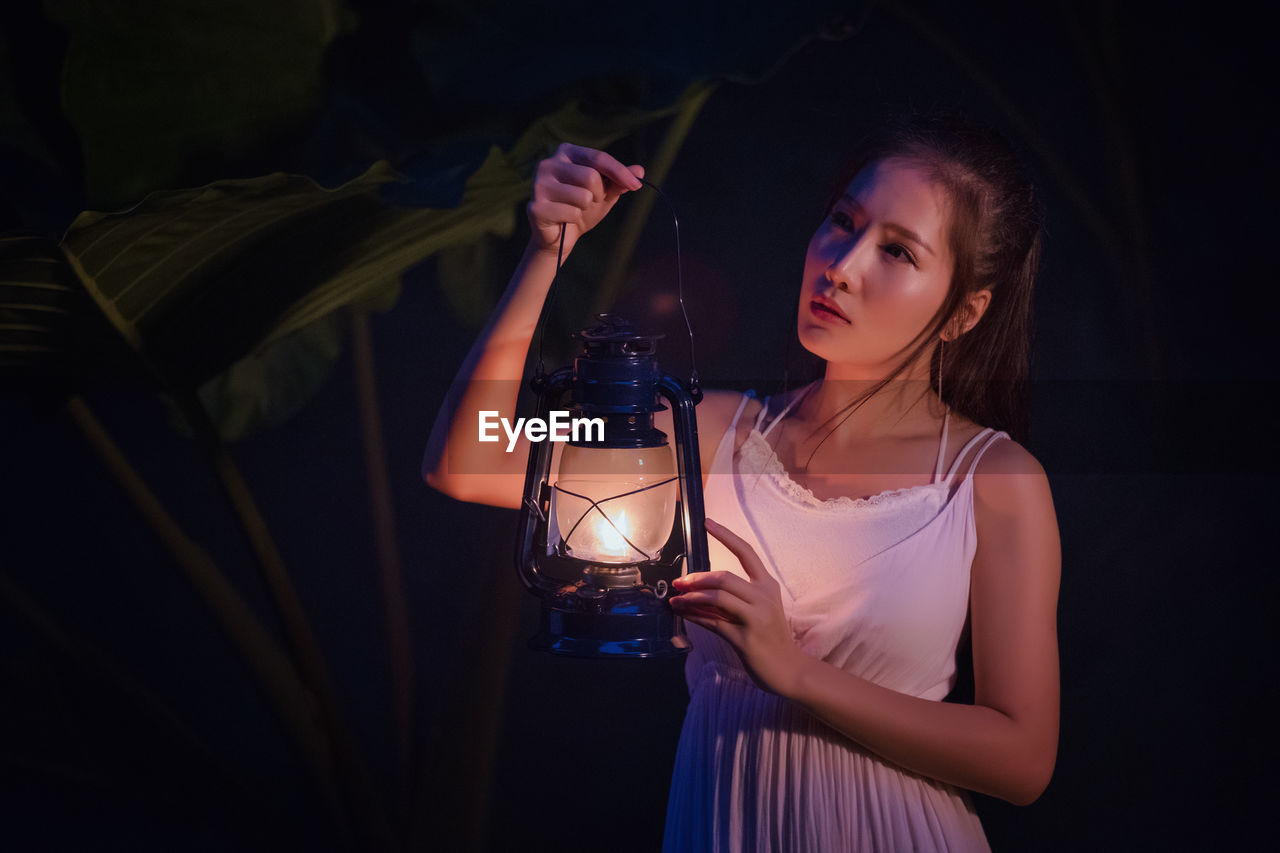 Woman holding illuminated lantern while standing outdoors at night