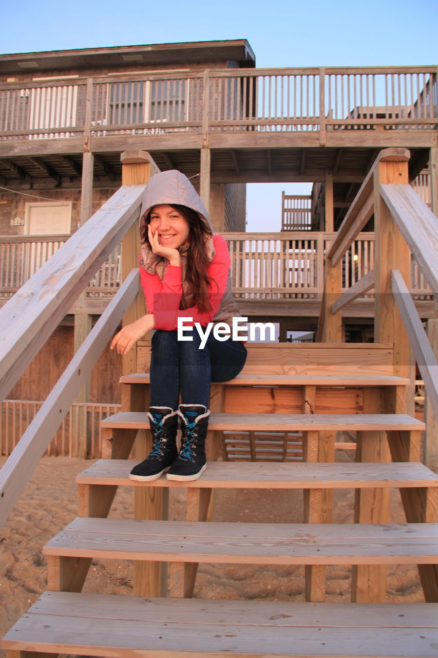 Full length portrait of smiling woman sitting on wooden steps at beach