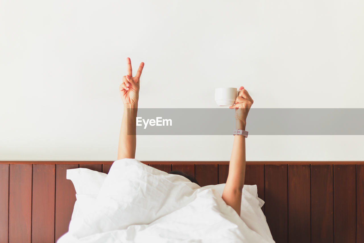 Female hand showing the funny sign and holding coffee cup behind blanket in the bed.