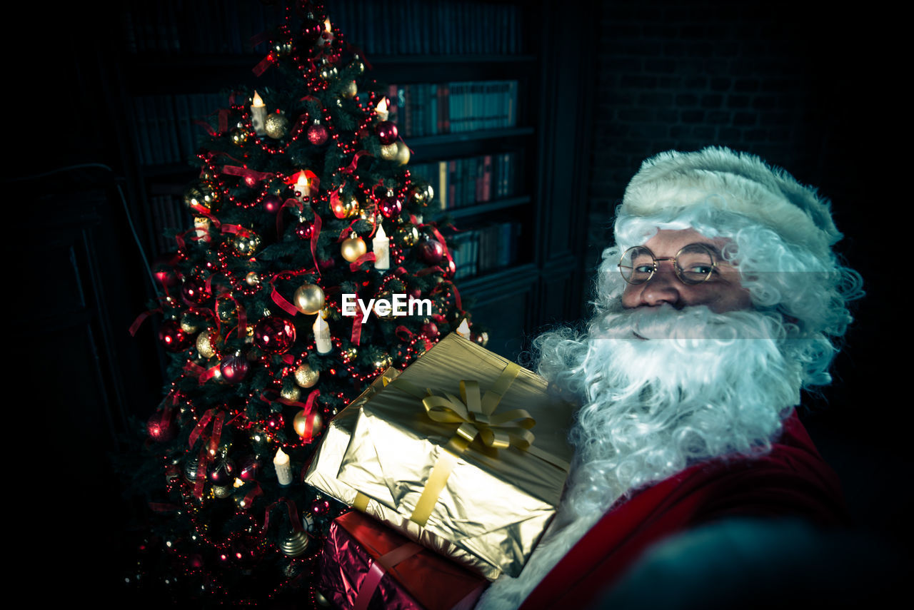 Portrait of man wearing santa claus costume holding christmas presents at home