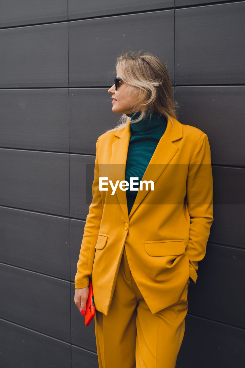 yellow, adult, one person, outerwear, women, jacket, standing, fashion, spring, clothing, coat, blond hair, business, looking, formal wear, young adult, hairstyle, three quarter length, sleeve, female, blazer, looking away, copy space, lifestyles, architecture, collar, business finance and industry, wall - building feature, smiling, businesswoman, outdoors, glasses