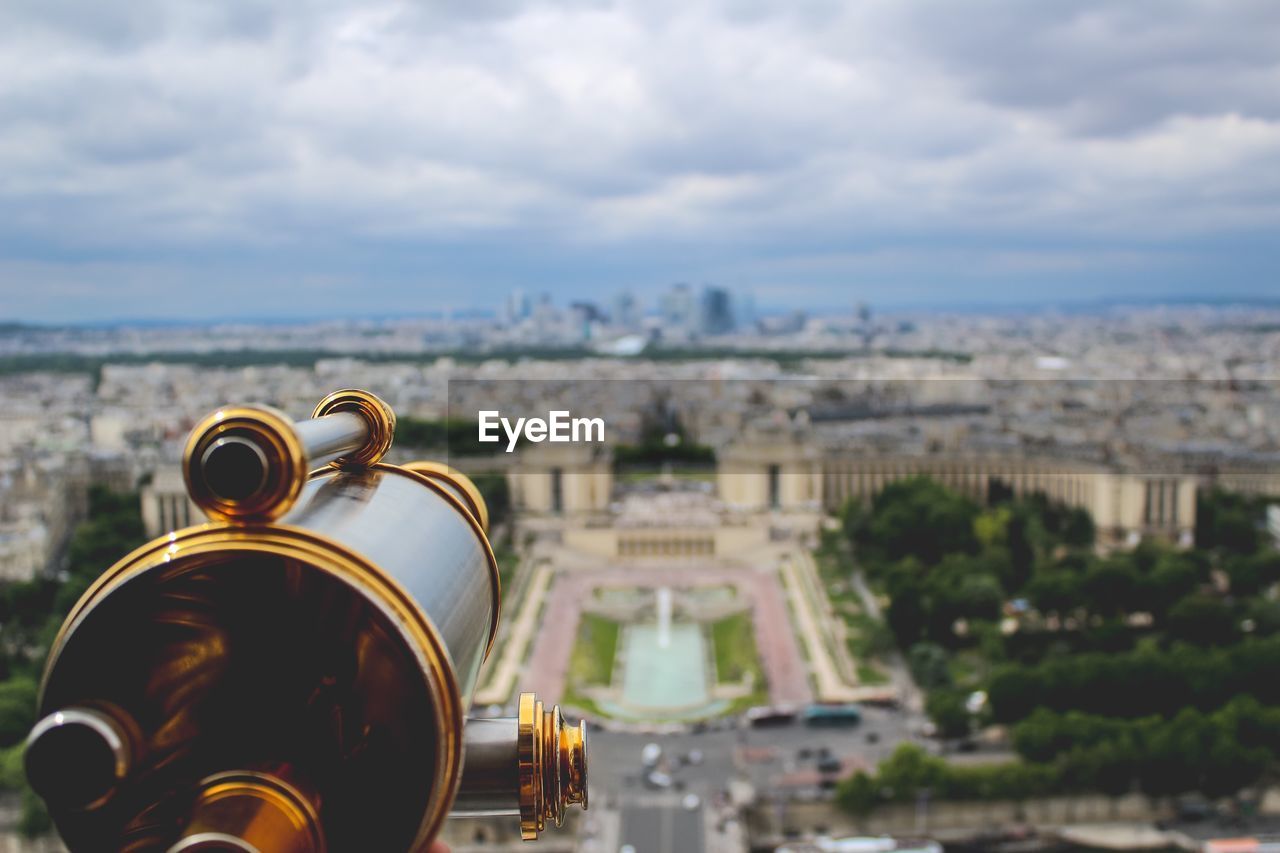 Close-up of coin-operated binoculars with buildings in background against cloudy sky