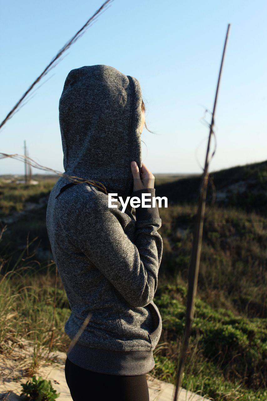 Side view of woman wearing hooded shirt standing on field against clear sky