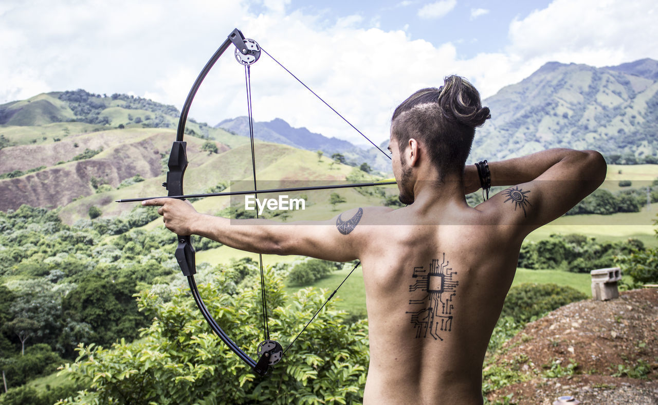Rear view of shirtless young man holding bow and arrow while standing on land