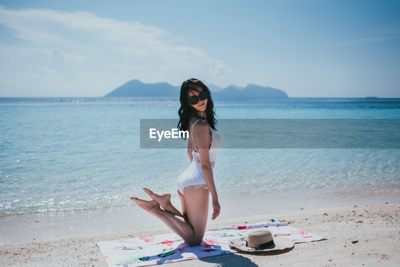 Side view of young woman wearing sunglasses while kneeling at beach against sky during sunny day