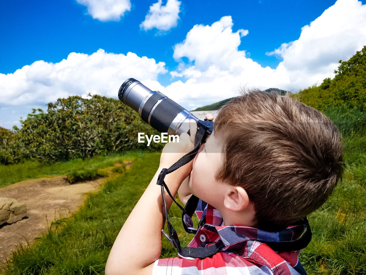 Side view of boy looking through binoculars while standing on field against sky