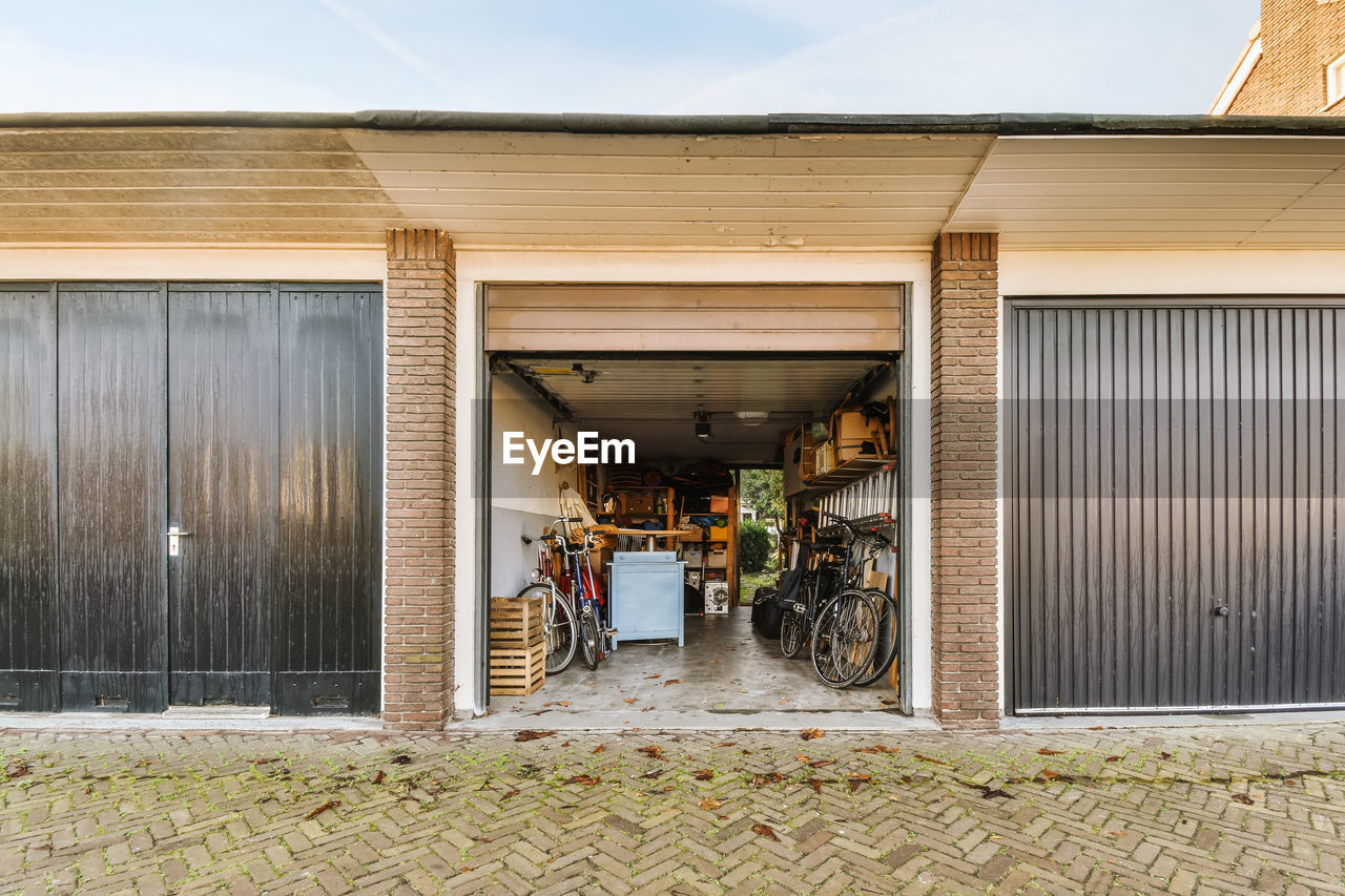 Opened metal garage with parked bicycles and wooden boxes with various equipment on shelves located in countryside on summer day