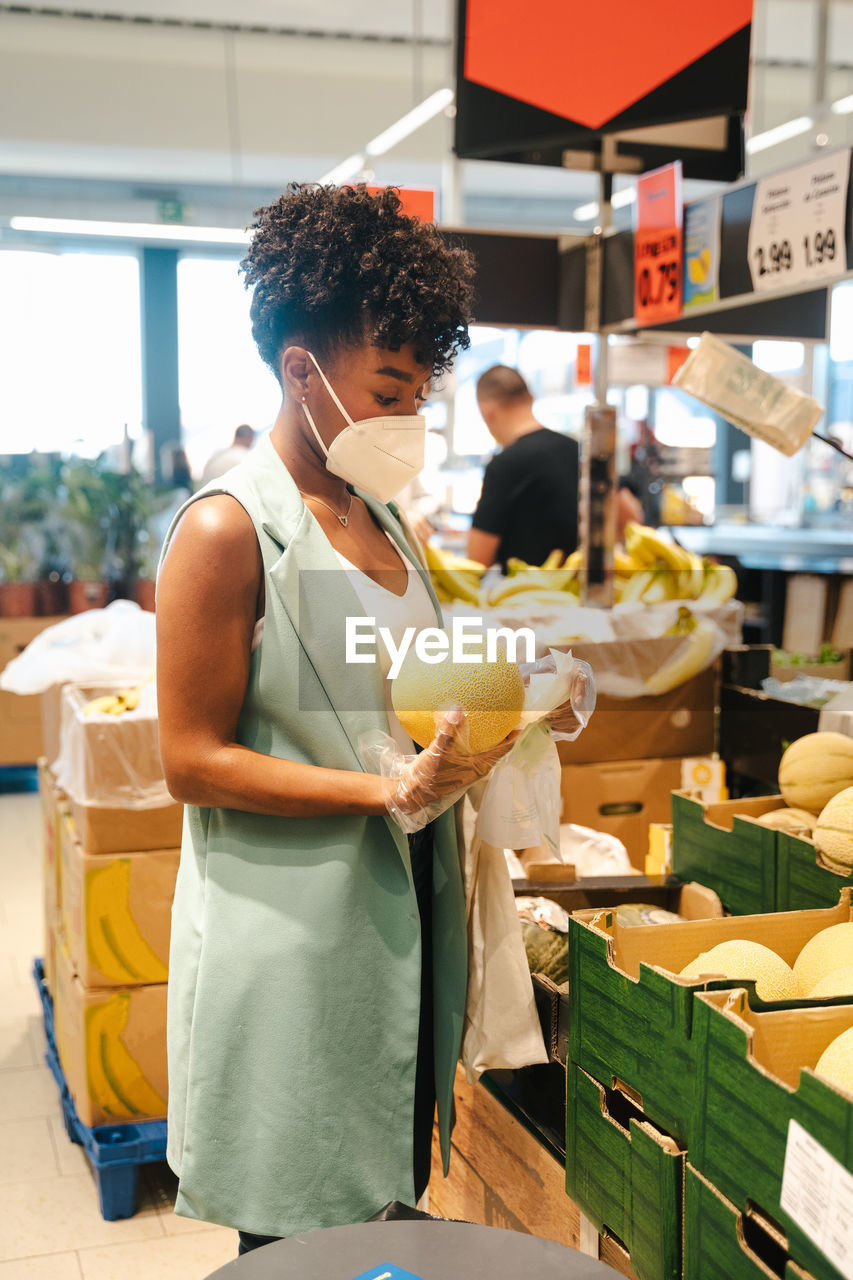 Side view of african american female customer in protective mask and gloves picking ripe watermelon from box while making purchases in supermarket during coronavirus pandemic