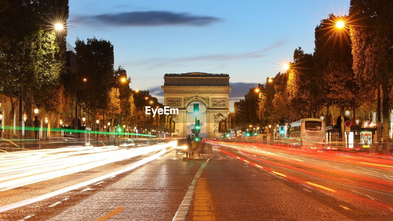 Light trails on city street by arc de triomphe against sky during dusk