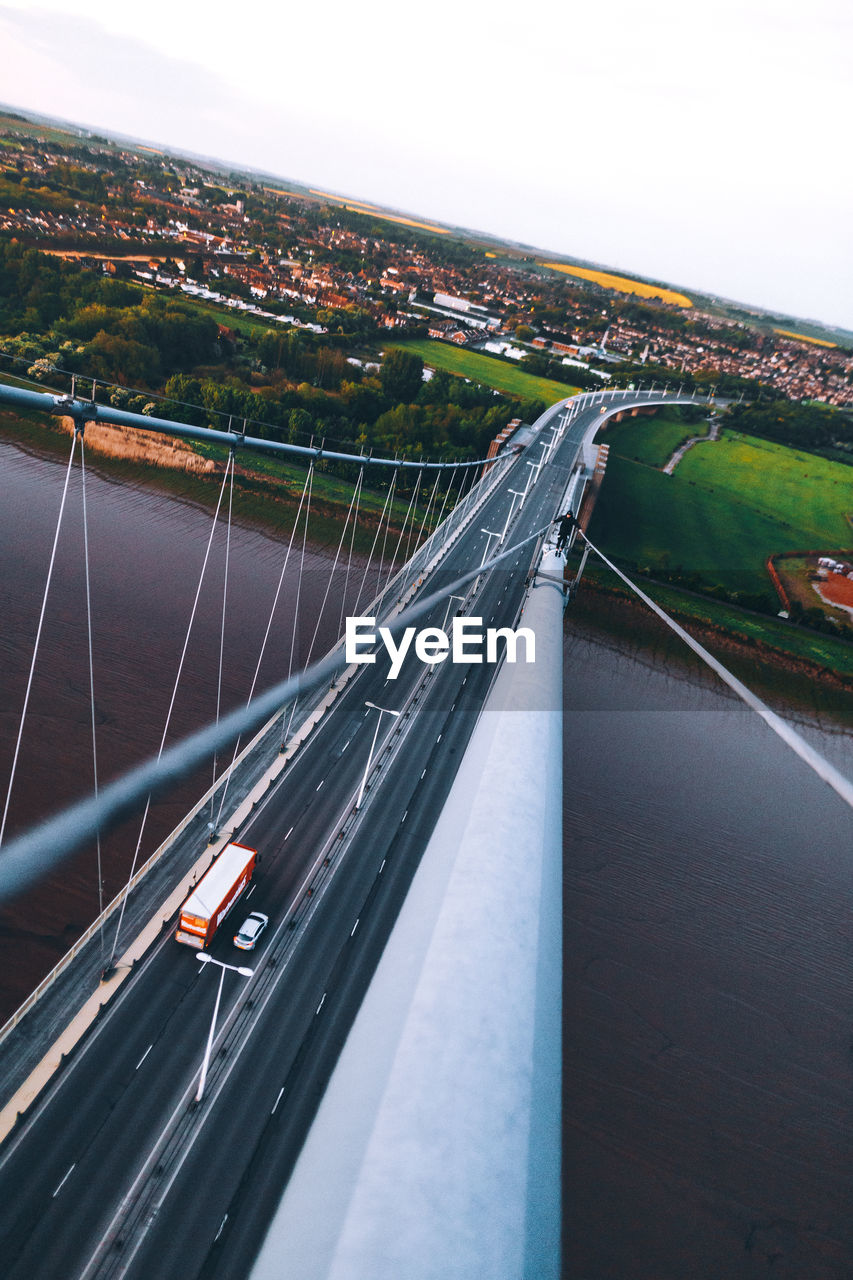 AERIAL VIEW OF SUSPENSION BRIDGE OVER RIVER IN CITY AGAINST SKY