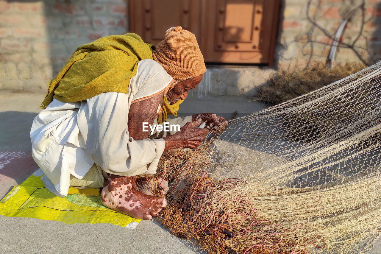 A very old but capable exemplary man of india making fishing nets in the sun 