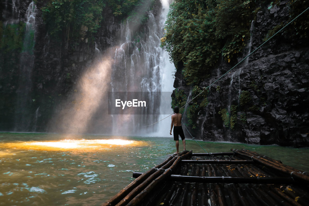 Rear view of shirtless man standing on wooden raft in river at forest