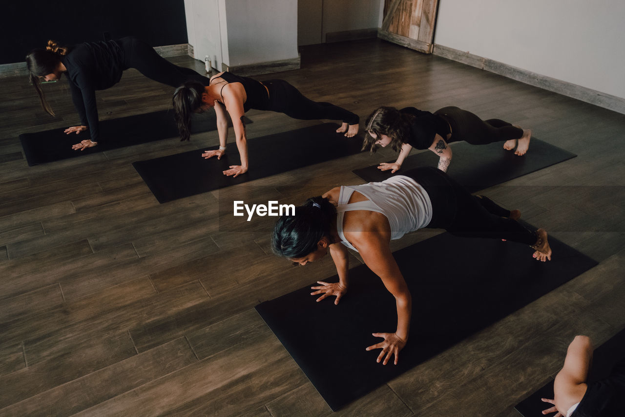 From above barefoot unrecognizable women in sportswear concentrating and doing plank exercise on sports mats on wooden floor against white walls of spacious hall