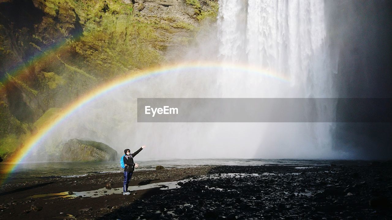 Mid adult woman standing by waterfall against rainbow