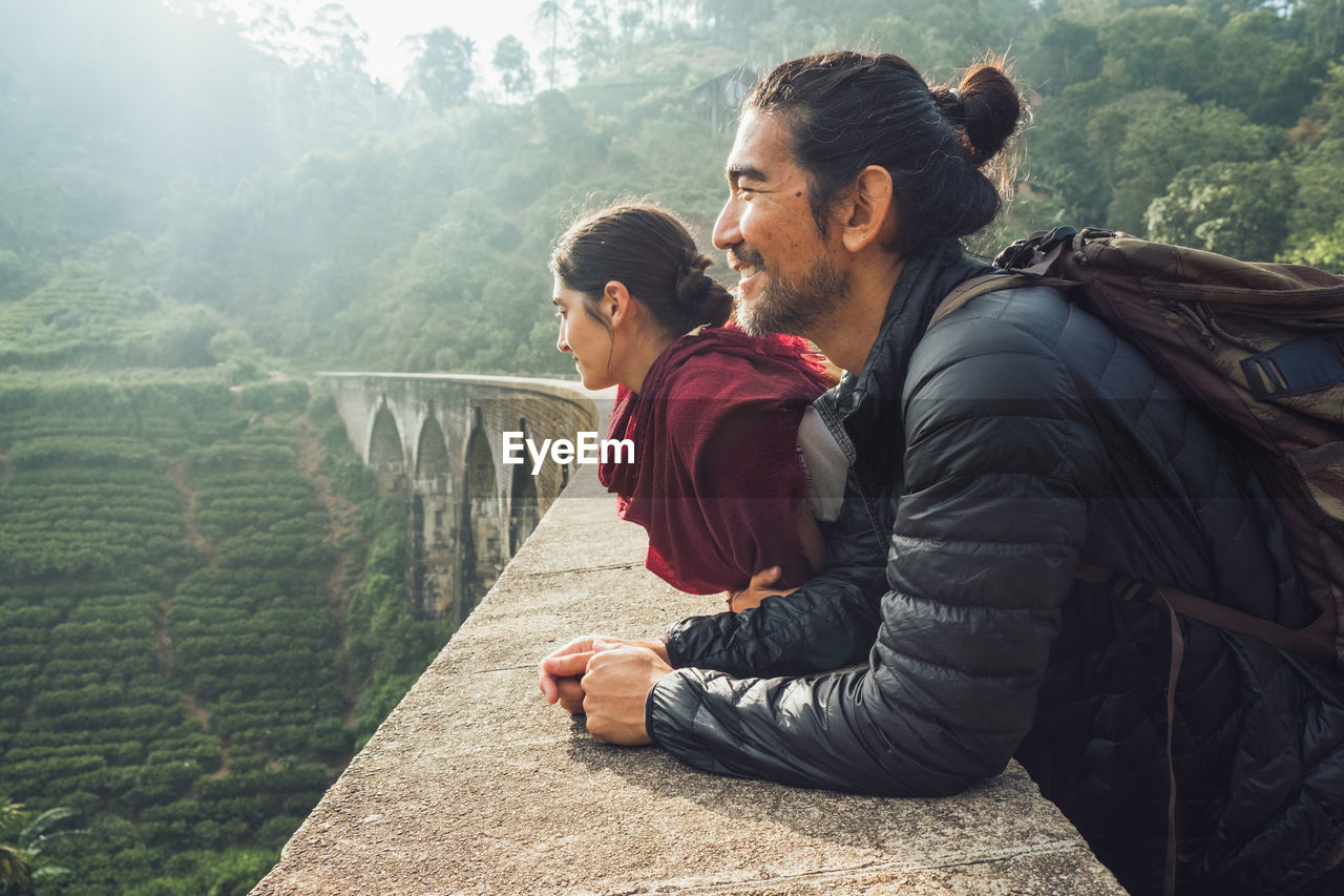 Side view of cheerful ethnic man and woman in activewear with backpacks standing contemplating views on aged stone nine arch bridge among green hills in sri lanka