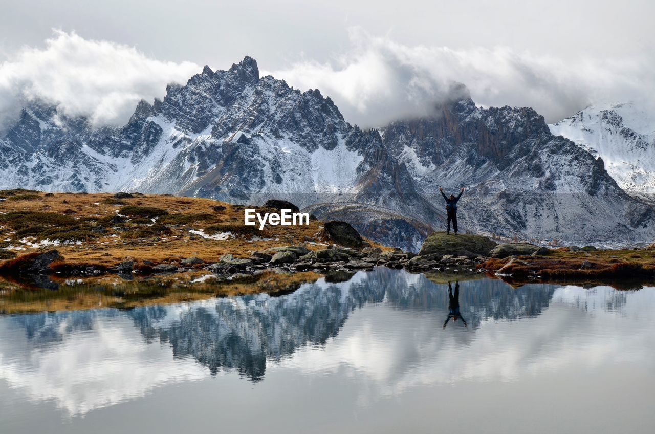 Man with arms raised reflecting on lake by mountains against cloudy sky