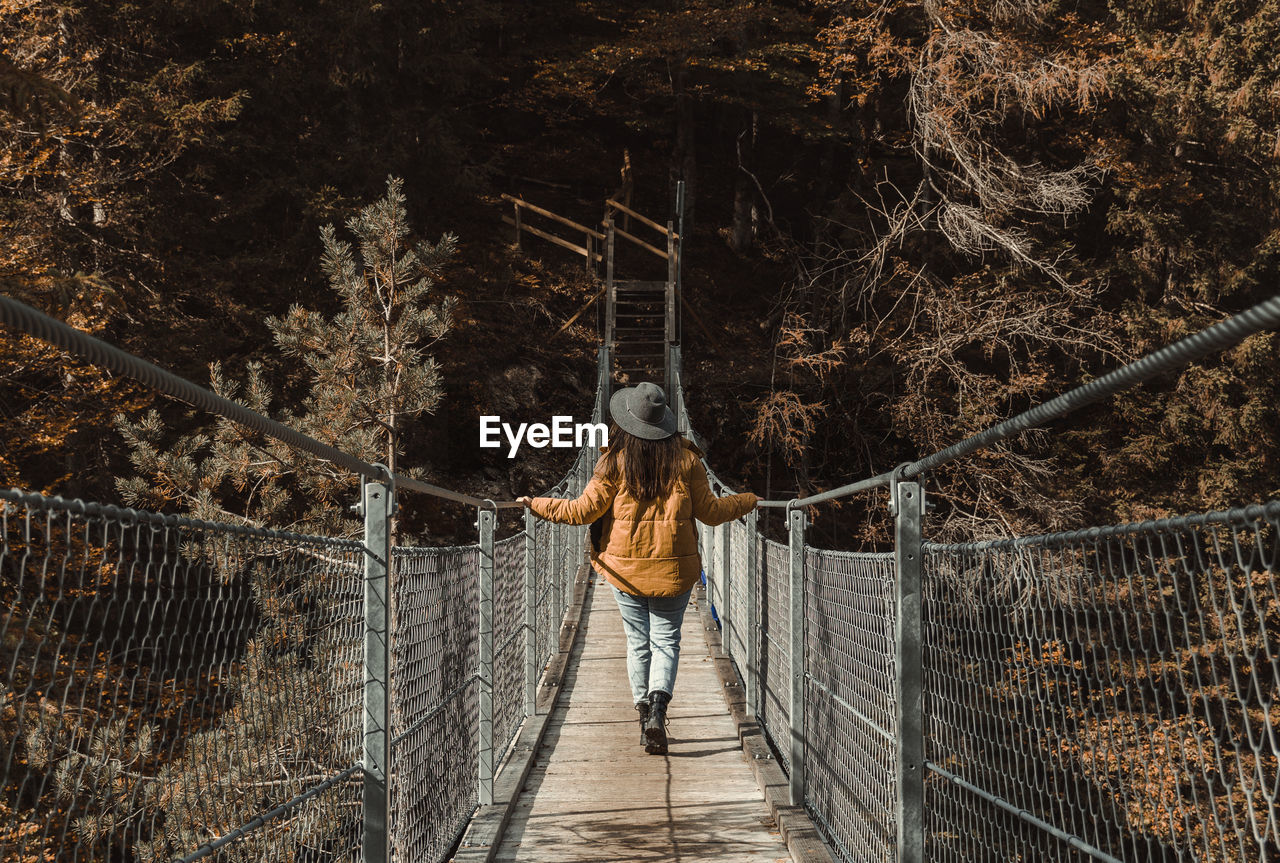 bridge, rope bridge, one person, full length, nature, leisure activity, rope, canopy walkway, tree, rear view, lifestyles, walking, suspension bridge, footbridge, casual clothing, the way forward, forest, architecture, railing, day, built structure, outdoors, land, adult, plant, standing, men, women, spring, young adult, travel, person, sunlight, tourism, transportation, motion