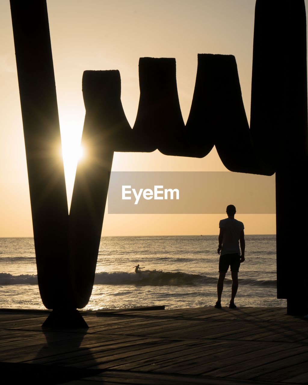 Silhouette man standing on beach against surfer during sunset