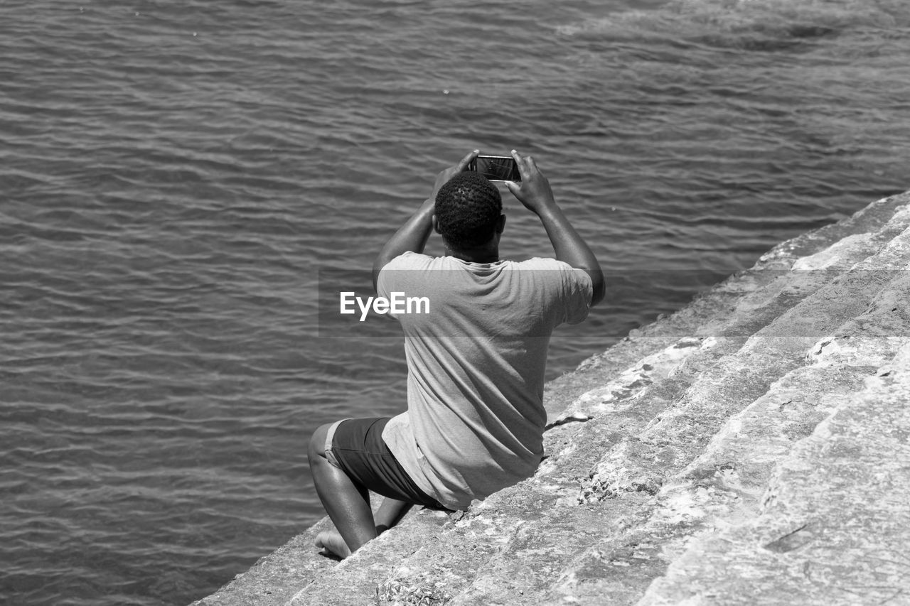 Rear view of man photographing through mobile phone while sitting on steps by sea