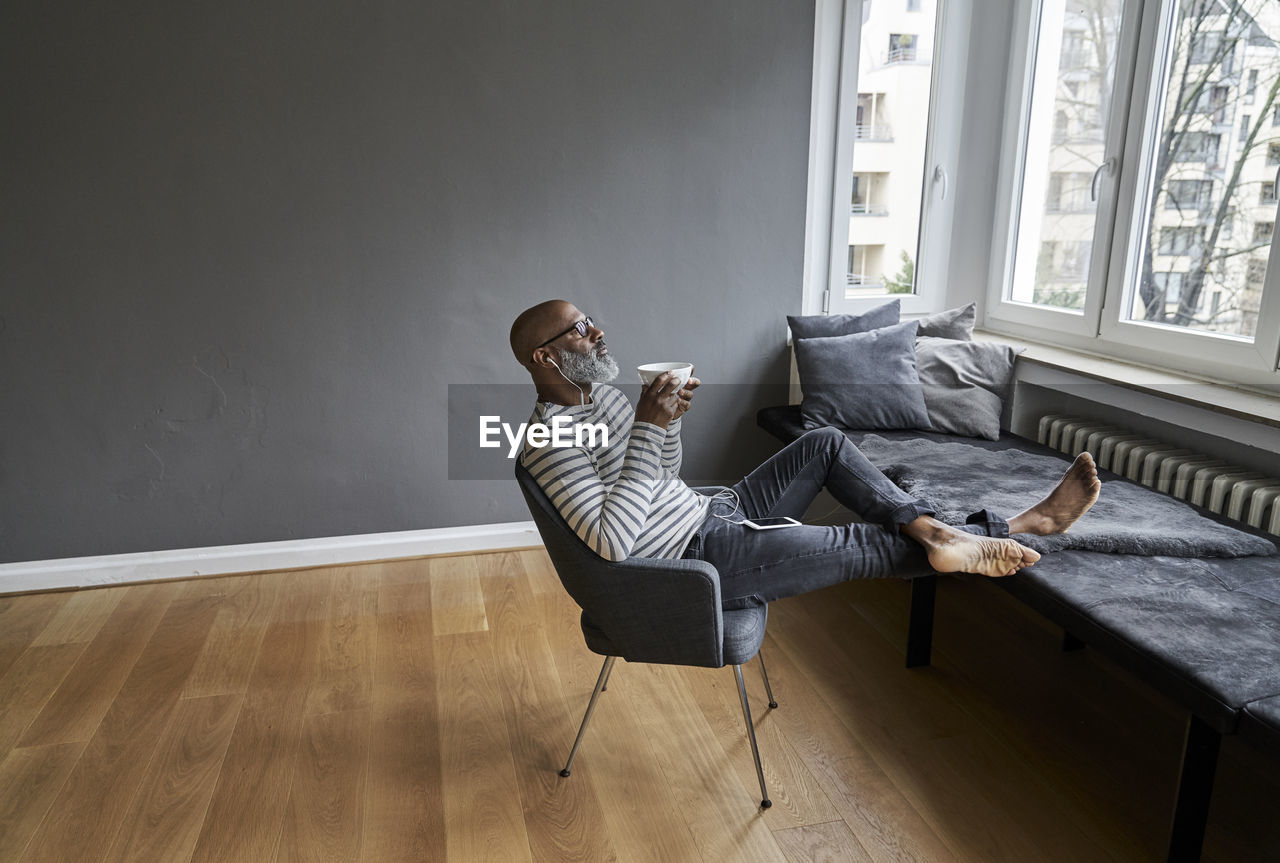 Matureman with earphones sitting at window, drinking coffee barefoot