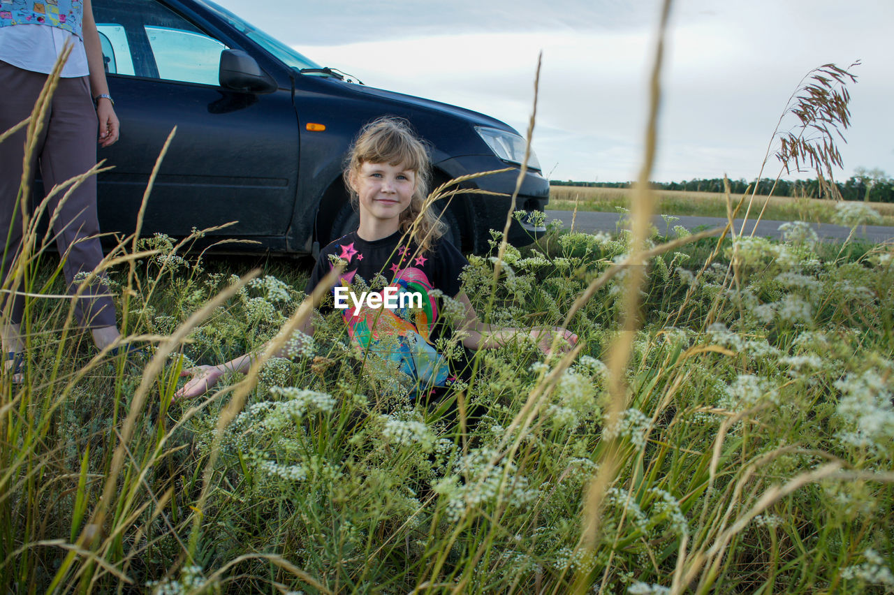 one person, nature, plant, land, women, child, grass, transportation, mode of transportation, childhood, sky, landscape, portrait, holiday, leisure activity, female, day, trip, vacation, looking at camera, rural scene, field, emotion, smiling, adult, front view, lifestyles, happiness, vehicle, adventure, travel, motor vehicle, outdoors, cloud, hairstyle, environment, flower, long hair, casual clothing, car, rural area, blond hair, person, enjoyment, growth, three quarter length