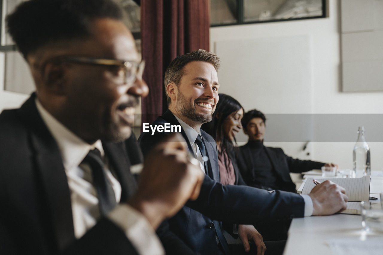 Smiling businessman looking away while sitting with colleagues during meeting at office