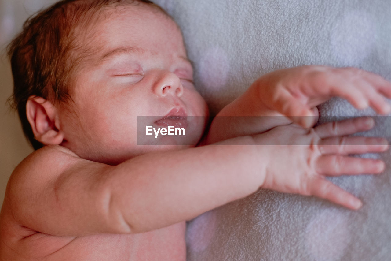 Calm cute infant with closed eyes covered with blanket resting in bed against blurred interior of light bedroom