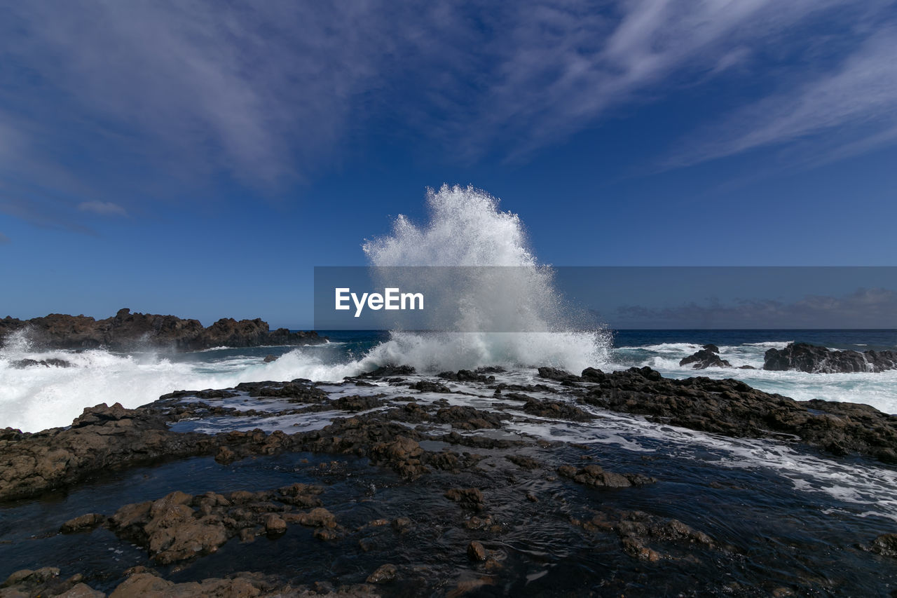 Waves splashing on rocks at shore against sky