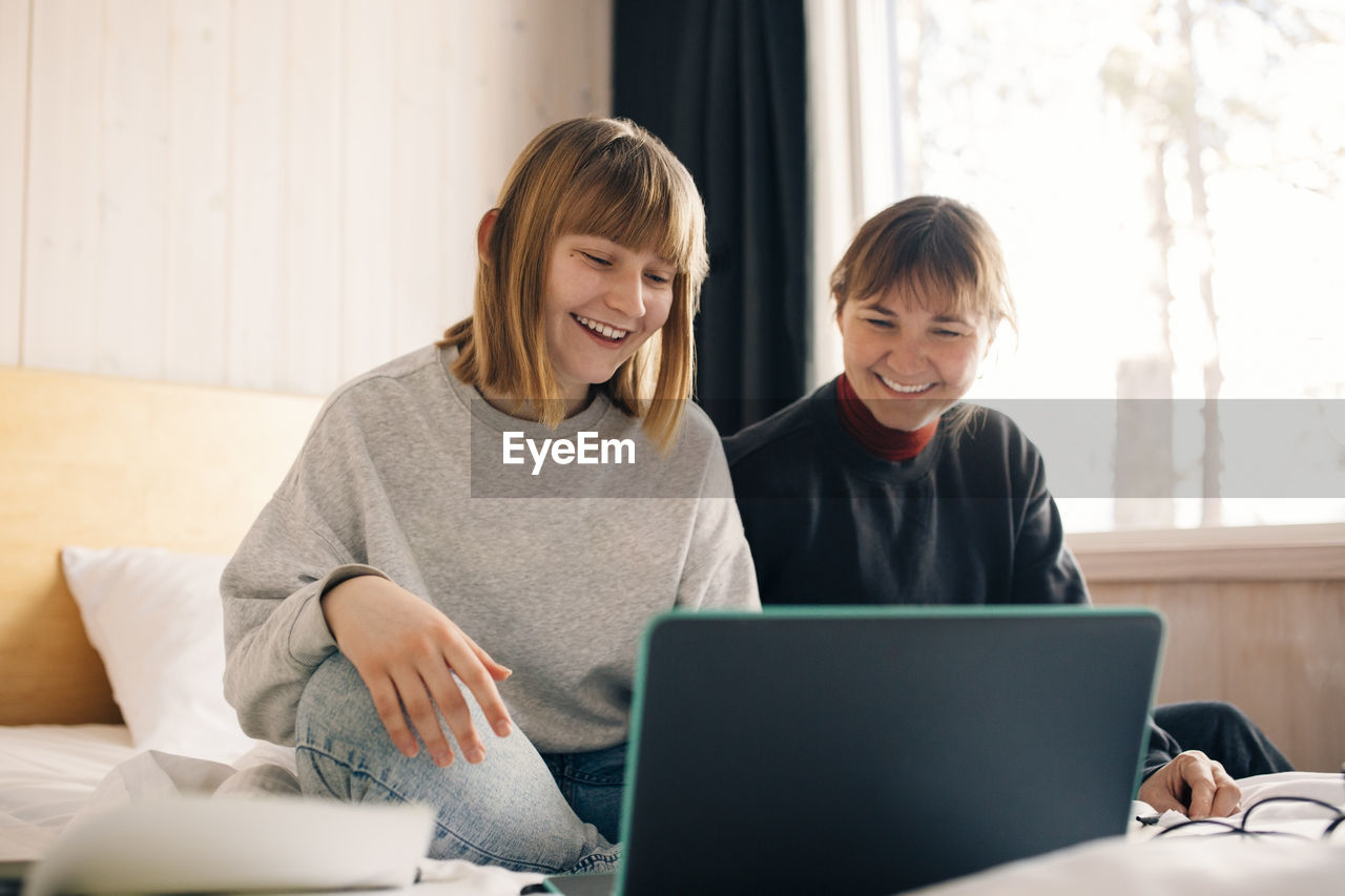 Smiling mother and daughter sitting with laptop while homeschooling in bedroom
